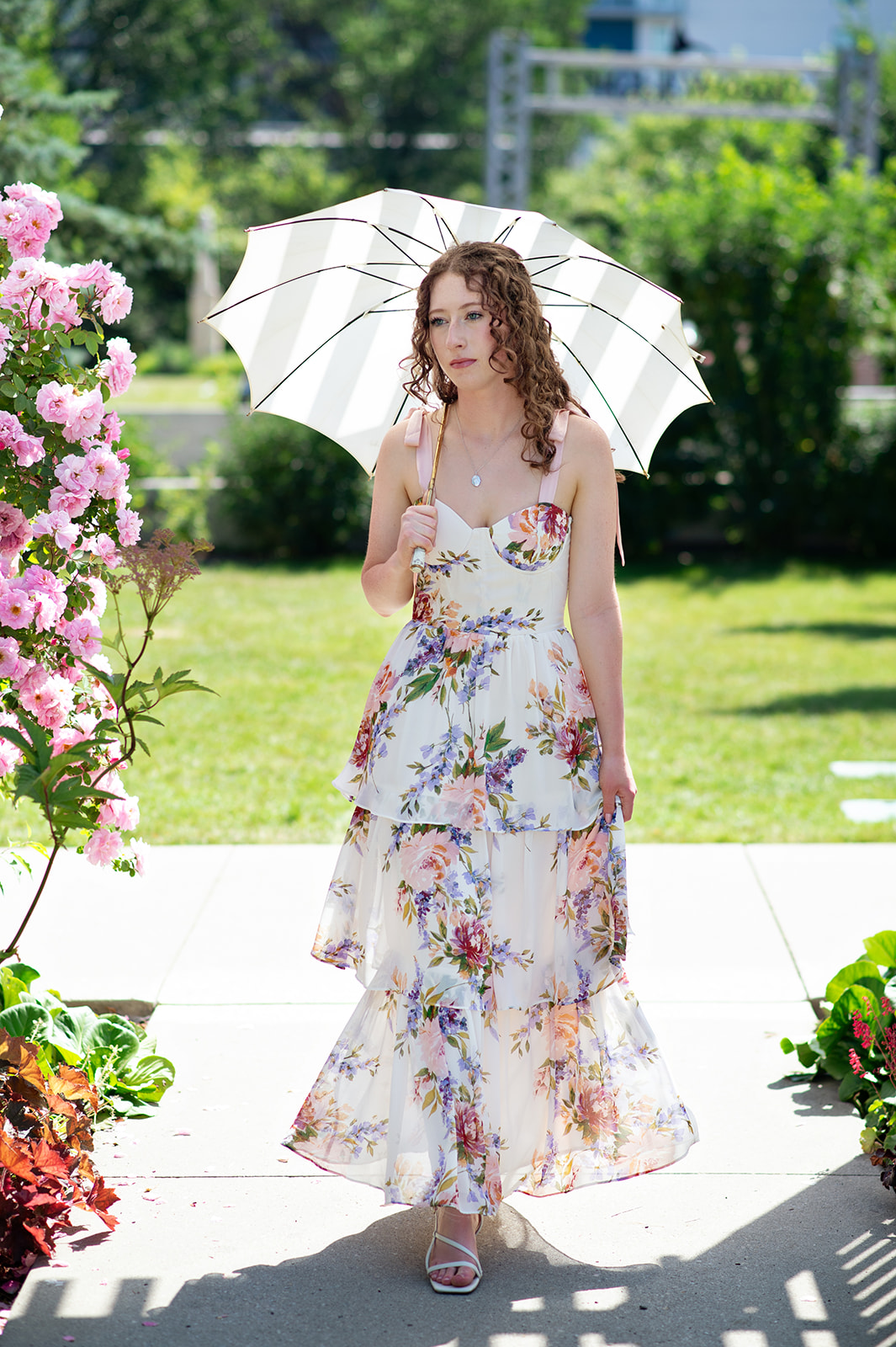 Woman in ruffled floral dress holding white umbrella walks through Deane House garden during summer wedding